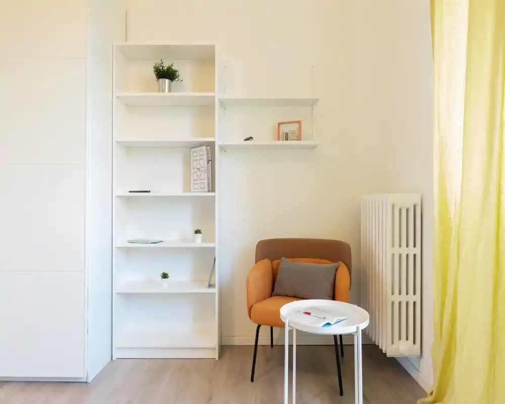 Rincón de salón luminoso y minimalista con un cómodo sillón naranja, una pequeña mesa redonda y estanterías blancas: ideal para leer o relajarse en un piso compacto.