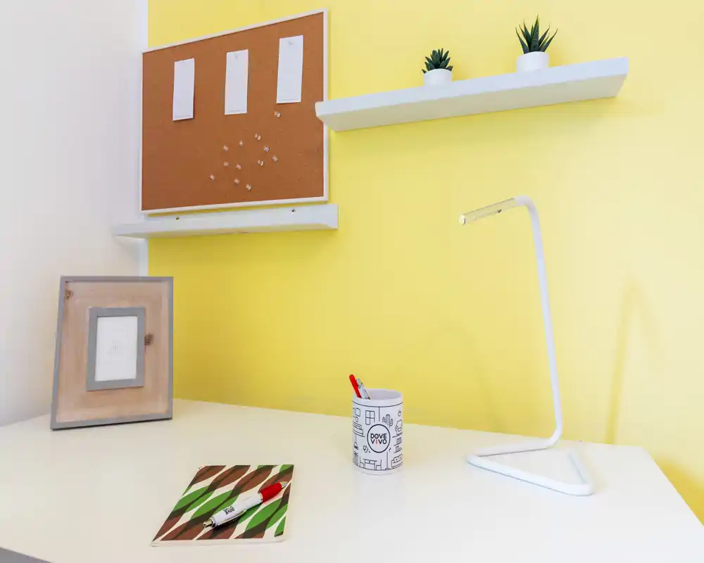 Esquina de escritorio luminosa y minimalista con lámpara blanca, corcho y pequeñas plantas: un espacio de trabajo ordenado ideal para teletrabajo.