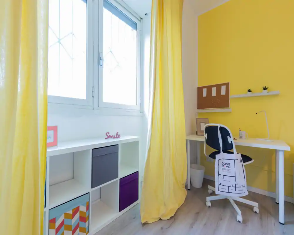 Rincón de oficina en casa luminoso y alegre con escritorio blanco, silla ergonómica y cubos de almacenaje coloridos junto a una gran ventana con cortinas amarillas.
