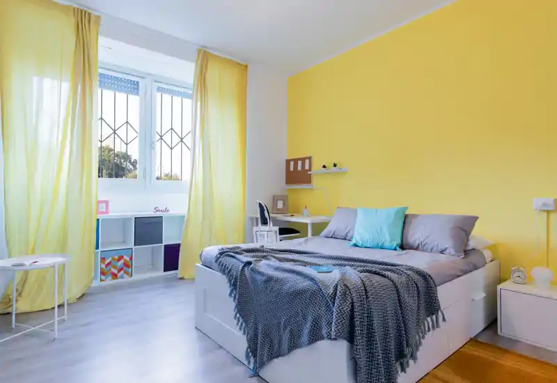 Dormitorio moderno y luminoso con cama doble con ropa de cama gris, una manta acogedora y un cojín turquesa, pared amarilla de acento y una gran ventana con cortinas amarillas que dejan entrar luz natural.