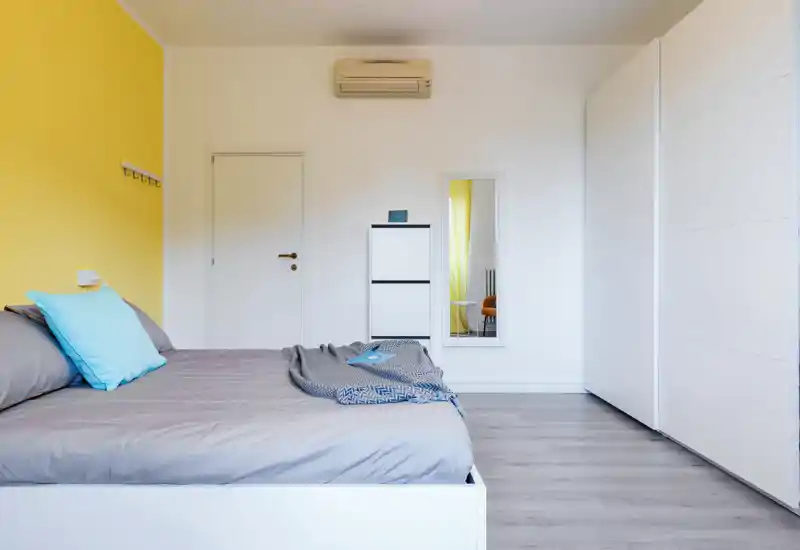Dormitorio moderno y luminoso con cama grande vestida en gris y cojines azules, armario blanco y espejo minimalista: ambiente tranquilo y aireado perfecto para anuncios.