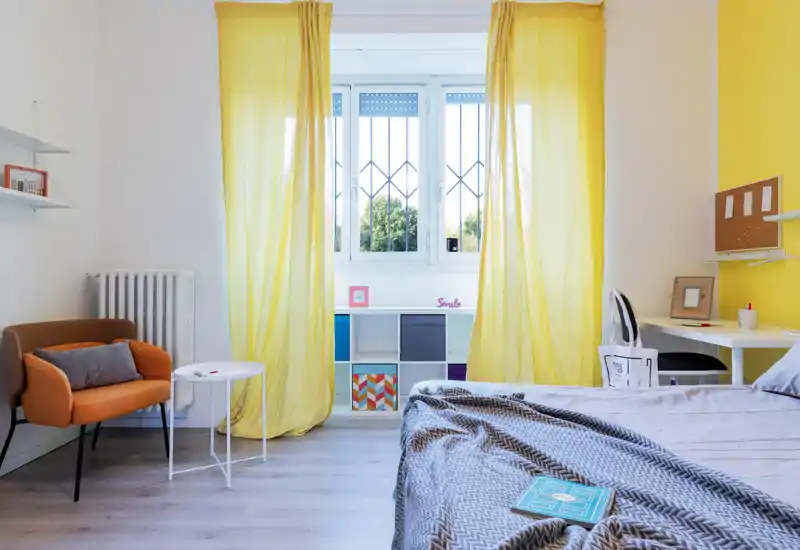 Dormitorio moderno y luminoso con cama en primer plano, cortinas amarillas alegres, un sillón cómodo y un pequeño escritorio — ideal para un alquiler soleado y acogedor.