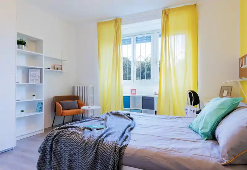 Dormitorio luminoso y bien decorado con cama doble, ropa de cama suave y una manta acogedora; gran ventana con cortinas amarillas que aporta mucha luz natural.