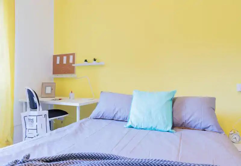 Dormitorio luminoso y ordenado con una cama grande vestida con ropa de cama suave, cojines en tonos pastel y un pequeño escritorio junto a la ventana — moderno y acogedor.