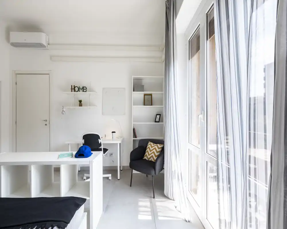 Dormitorio moderno y luminoso con vista parcial de la cama, escritorio, estanterías y un sillón junto a grandes ventanas que aportan mucha luz natural.