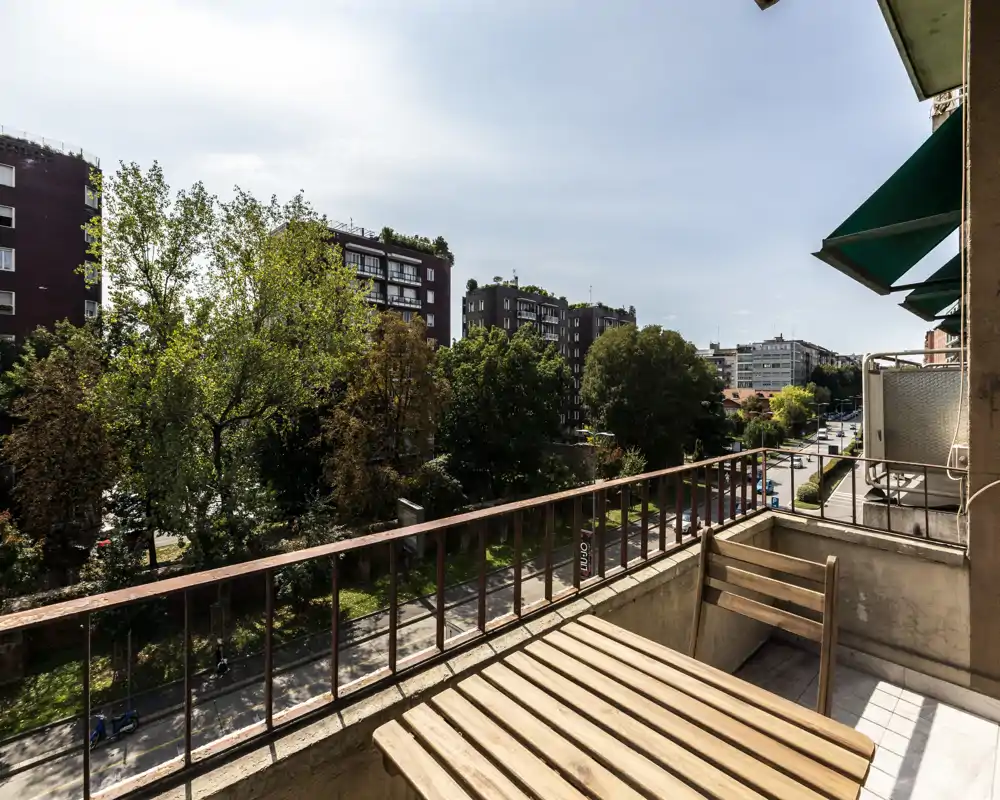 Balcón soleado con mesa bistró de madera y silla con vistas a una calle arbolada y edificios — ideal para el café matutino.