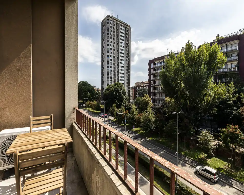 Balcón soleado con pequeña mesa y sillas de madera con vistas a una calle arbolada y edificios altos, ideal para tomar un café por la mañana y disfrutar de la vista urbana.
