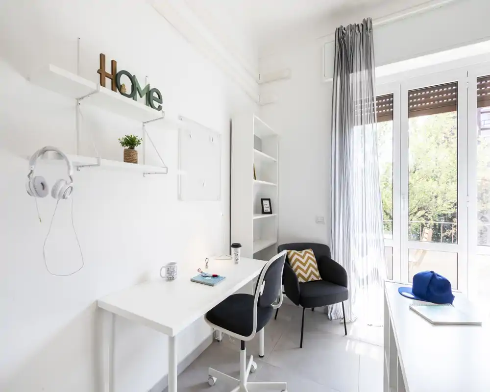 Espacio de trabajo moderno y luminoso con escritorio blanco, silla giratoria y un sillón acogedor junto a una gran ventana que aporta abundante luz natural.