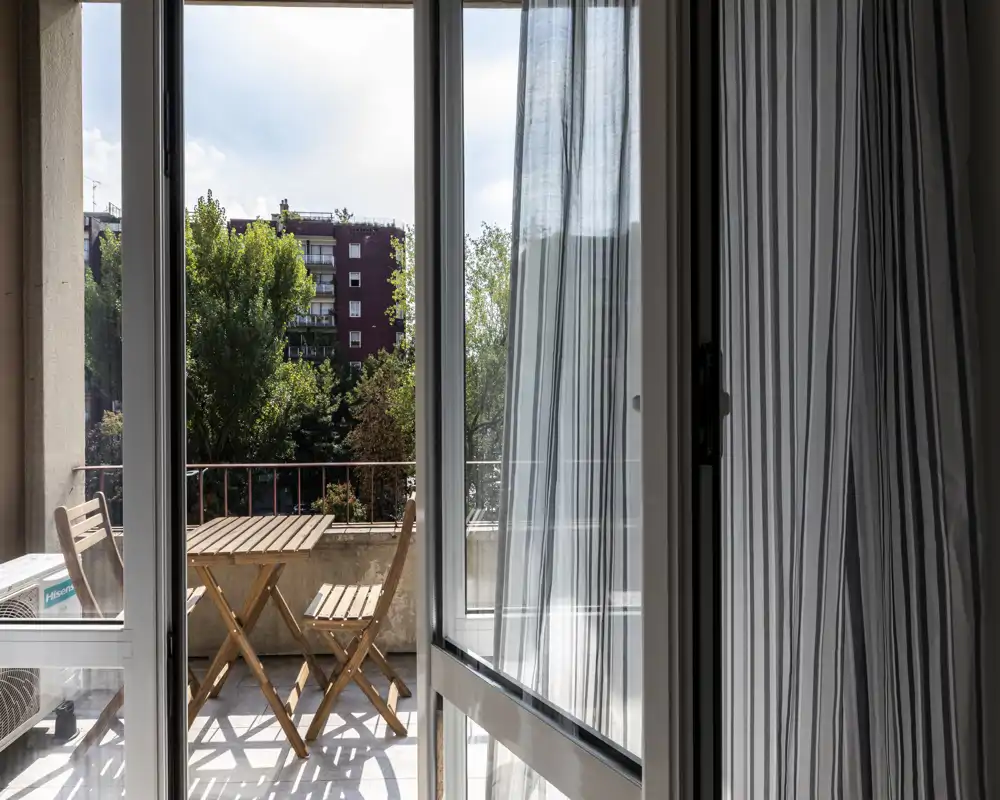 Balcón soleado con una pequeña mesa de madera y dos sillas plegables, visto a través de puertas correderas; agradable vista urbana y luz natural.