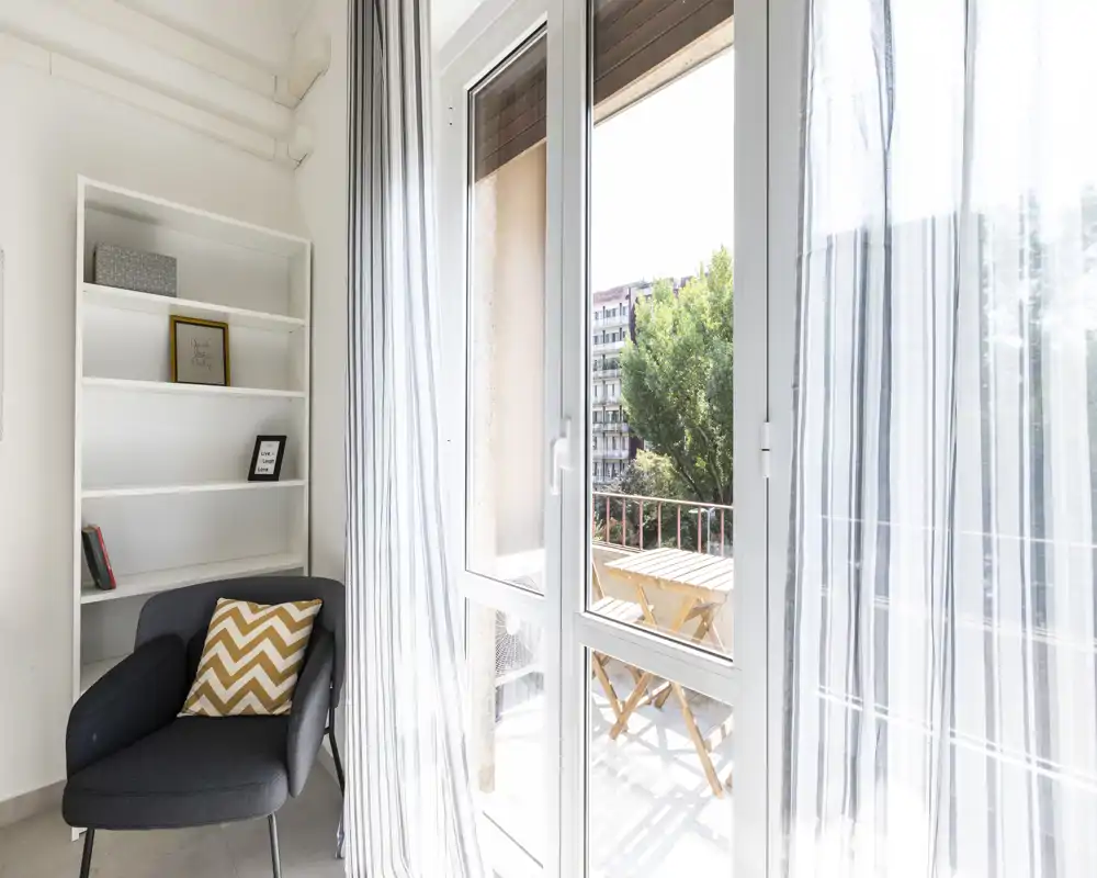 Rincón de salón luminoso y aireado con un sillón cómodo, cortinas translucidas a rayas y un balcón con mesa exterior, ideal para relajarse con luz natural.