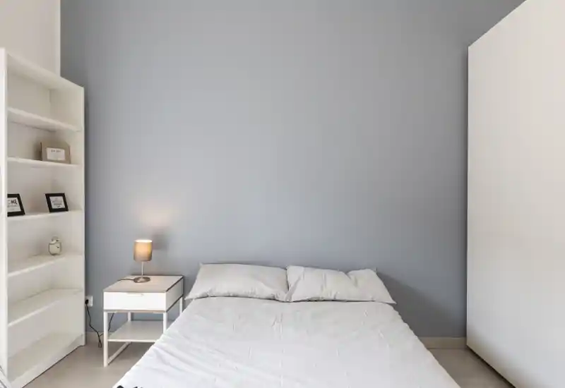 Dormitorio minimalista con cama doble frente a una pared azul suave, mesita con lámpara, estantería blanca y armario — luminoso, limpio y de estilo sencillo para una atmósfera tranquila.