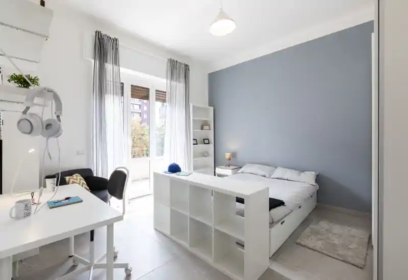 Dormitorio moderno y luminoso con cama doble frente a una pared azul apagada, estantería integrada y balcón que aporta mucha luz. Estilo minimalista limpio con lámpara de noche, alfombra y estantería baja que separa la zona de descanso del espacio de trabajo.