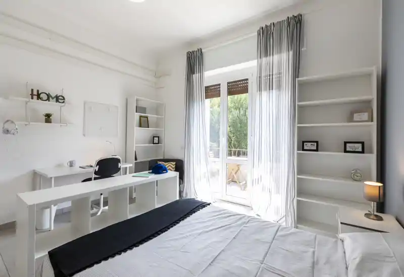 Dormitorio moderno y luminoso con cama doble en primer plano, escritorio y estanterías, y una gran puerta que da al balcón aportando abundante luz natural.