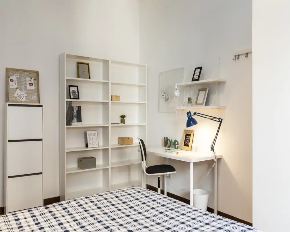 Dormitorio luminoso y bien presentado con cama estampada en primer plano, estantería blanca ordenada y un escritorio compacto con lámpara azul — ideal para una persona o estudiantes.