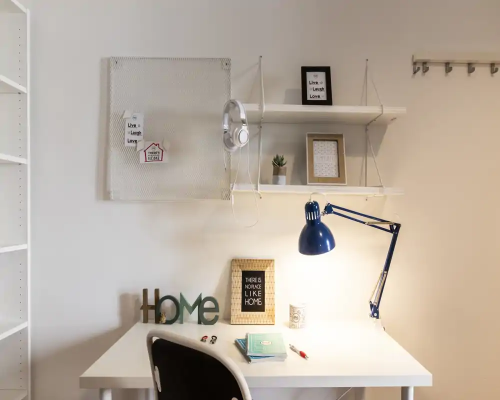 Rincón de oficina en casa luminoso y decorado con escritorio blanco, lámpara azul ajustable y estanterías decorativas — un espacio de trabajo ordenado para una persona.