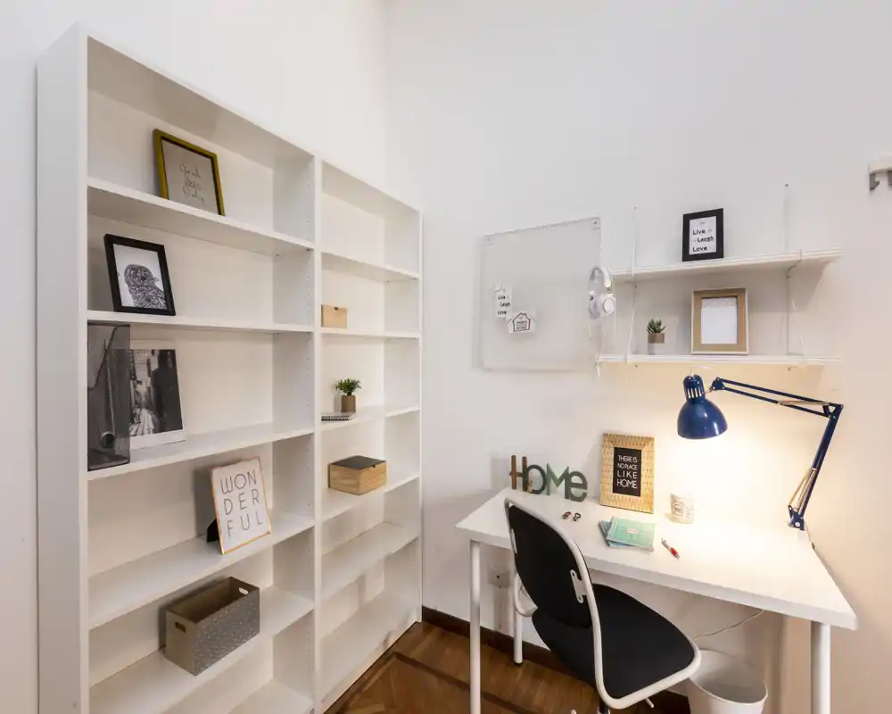 Esquina de oficina en casa luminosa y minimalista con escritorio blanco, lámpara de escritorio azul regulable, estanterías y detalles decorativos — ideal para trabajar o estudiar.