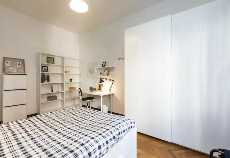 Dormitorio moderno y luminoso con una cama a cuadros grande en primer plano, armario blanco, estanterías y un pequeño escritorio con lámpara de trabajo — minimalista, limpio y acogedor.