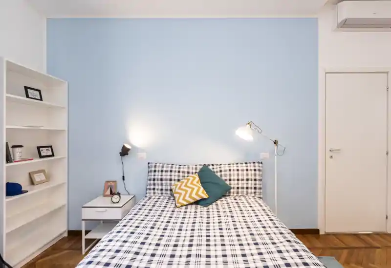 Dormitorio luminoso y bien arreglado con cama doble con ropa de cuadros, cojines decorativos y lámparas de mesita frente a una pared azul claro.