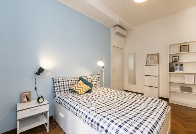 Dormitorio moderno y luminoso con cama doble con ropa de cuadros, mesita y estantería — acogedor y bien preparado para un anuncio.