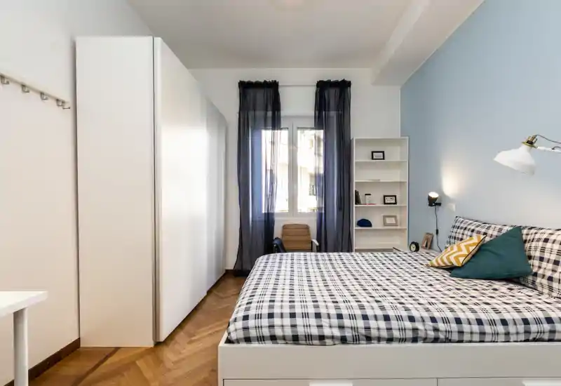 Dormitorio doble luminoso y bien decorado con cama grande de cuadros, armario blanco, estantería y luz natural desde la ventana — acogedor y moderno.