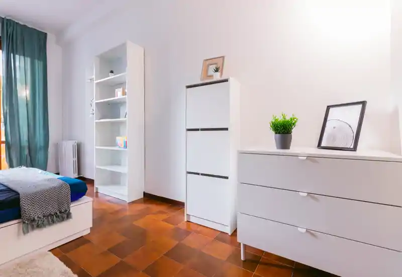 Bright, minimal bedroom with a neatly made bed, white storage units and warm tiled flooring, lit by natural light from the window.