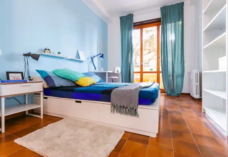 Bright, neatly staged bedroom with a full bed, bedside table, desk by a large window and pleasant natural light — ideal for rental photos.