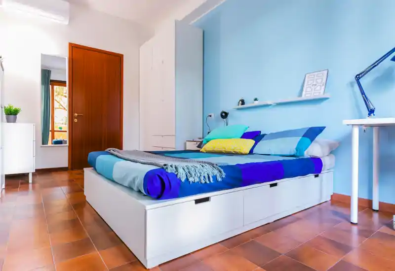 Bright, well-styled bedroom with a large white platform bed, layered blue bedding and colorful cushions; desk and storage complete the modern, airy layout.