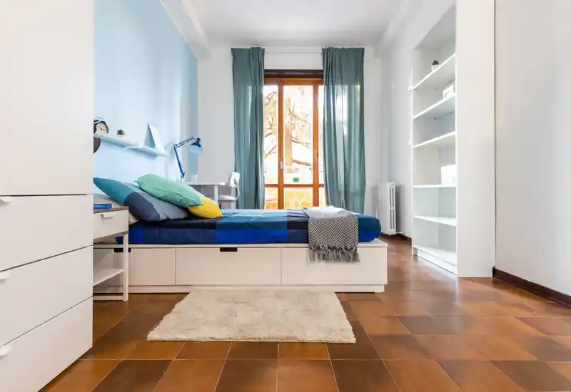 Bright, neatly styled bedroom with a low platform bed, layered cushions and a window letting in natural light. Built-in storage under the bed, a small desk by the window and a soft rug complete the cozy layout.