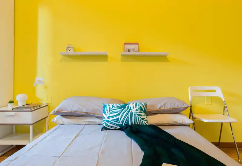Camera da letto luminosa e moderna con un ampio letto su una parete gialla vivace, cuscini decorativi e una coperta accogliente; comodino e sedia completano l'ambiente invitante.