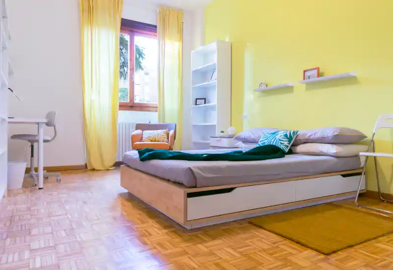 Camera da letto luminosa e moderna con letto basso in legno, biancheria grigia e accenti gialli caldi. La luce naturale entra dalla finestra con tende gialle; scaffali e una piccola scrivania rendono lo spazio funzionale e accogliente.
