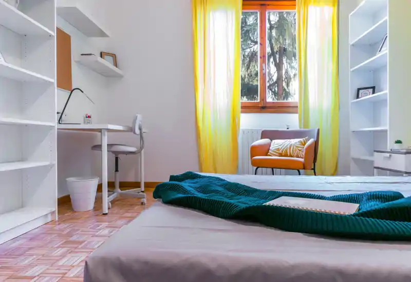 Camera da letto luminosa e ben allestita con un letto basso in primo piano, una coperta teal accogliente e una poltrona comoda vicino alla finestra che lascia entrare luce naturale.