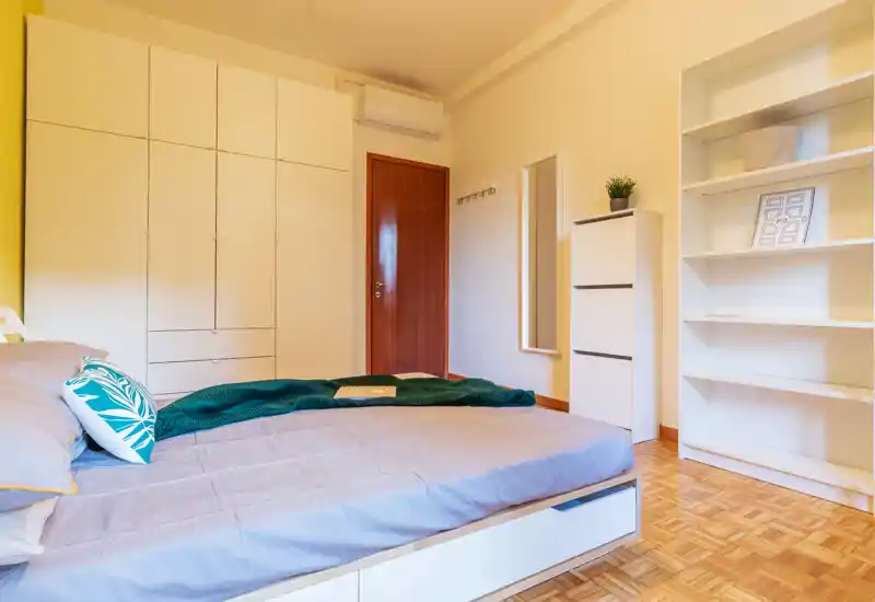 Camera da letto luminosa e ordinata con letto basso, armadio a muro e scaffalatura aperta; pavimento in legno e arredi moderni creano un’atmosfera accogliente.
