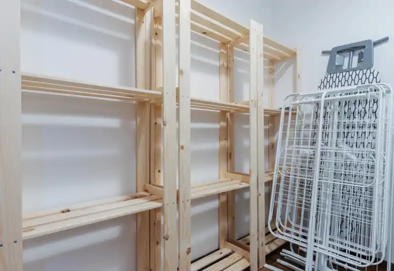 Pequeño trastero/área de servicio con estanterías de madera y tablas de planchar plegadas; espacio luminoso y funcional para organizar objetos del hogar.