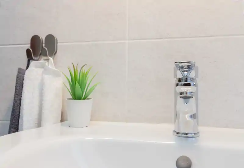Primer plano del área del lavabo de un baño moderno con grifo cromado, seno blanco, pared alicatada y toallas colgadas junto a una pequeña planta en maceta.