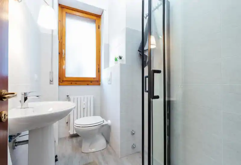 Baño compacto y moderno con lavabo de pedestal, inodoro y cabina de ducha de cristal, luz natural brillante desde una ventana translúcida.