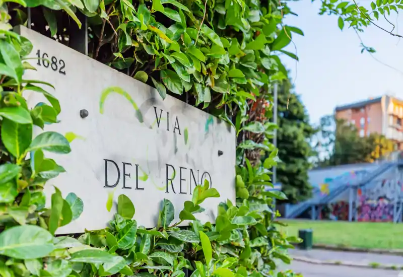 Vista desde la calle de una placa de mármol « Via Del Reno » enmarcada por follaje verde, con una zona ajardinada y una rampa con graffitis en el fondo.
