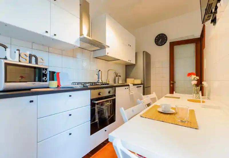 Cocina moderna y luminosa con muebles blancos, cocina de gas, horno y mesa para desayunos para dos — ambientada con manteles individuales y flores frescas para mayor calidez.