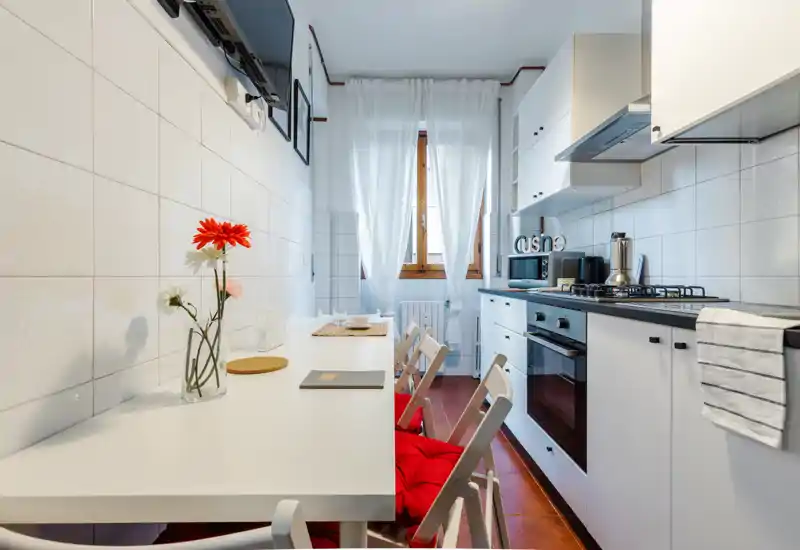 Cocina moderna y luminosa con muebles blancos, placa de gas, horno y una barra con sillas plegables con cojines rojos — ideal para apartamentos compactos.