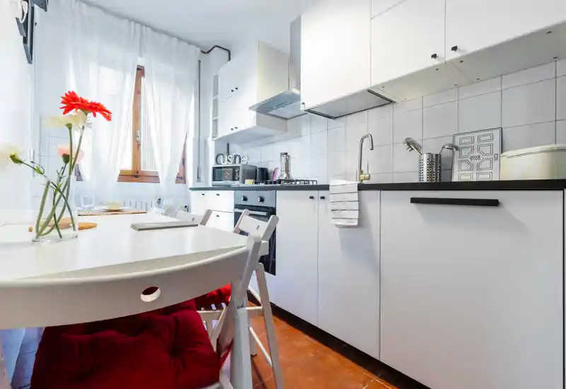 Cocina moderna y luminosa con armarios blancos, placa de gas y mesa junto a la ventana; decorada con flores frescas para una sensación acogedora.