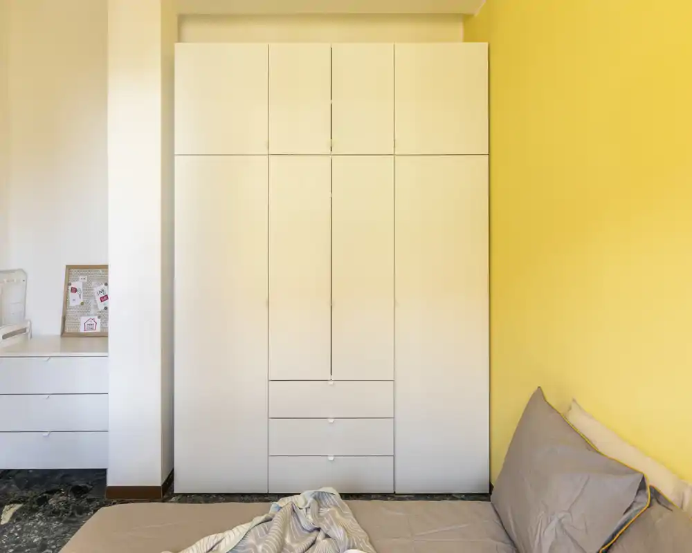 Rincón de dormitorio luminoso y ordenado con un gran armario blanco y una cama doble parcialmente visible, ropa de cama neutra y una pared amarilla cálida.