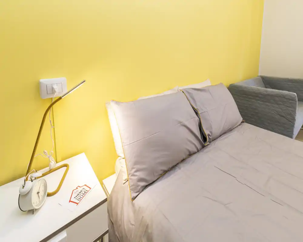 Dormitorio moderno y luminoso con cama doble bien hecha, ropa de cama gris suave, mesita con lámpara y despertador, y una pared amarilla de acento.