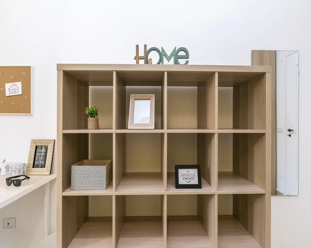 Rincón de salón luminoso y minimalista con una estantería modular de madera utilizada como separador, marcos decorativos y una pequeña planta que aporta calidez.