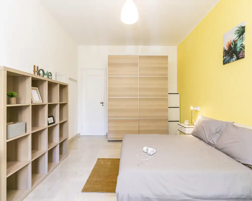 Dormitorio moderno y luminoso con gran cama doble, mesita de noche y armario de madera. Pared de acento amarilla y estantería ordenada, perfecto para un anuncio de alquiler.