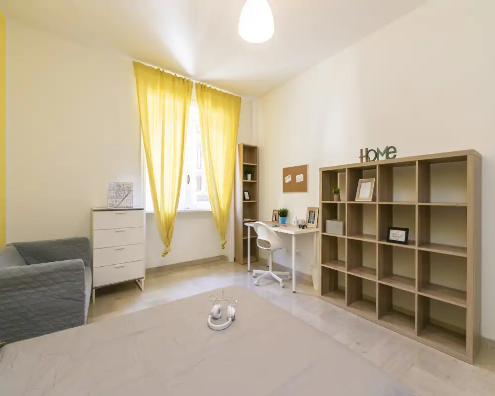 Dormitorio luminoso y de mobiliario sencillo con cama en primer plano, escritorio junto a la ventana y estantería abierta para almacenamiento.