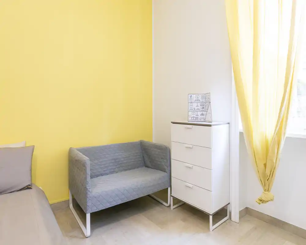 Rincón de dormitorio con cama parcialmente visible a la izquierda, un pequeño sofá gris y una cómoda blanca junto a una ventana luminosa con cortinas amarillas — aireado, moderno y bien iluminado.