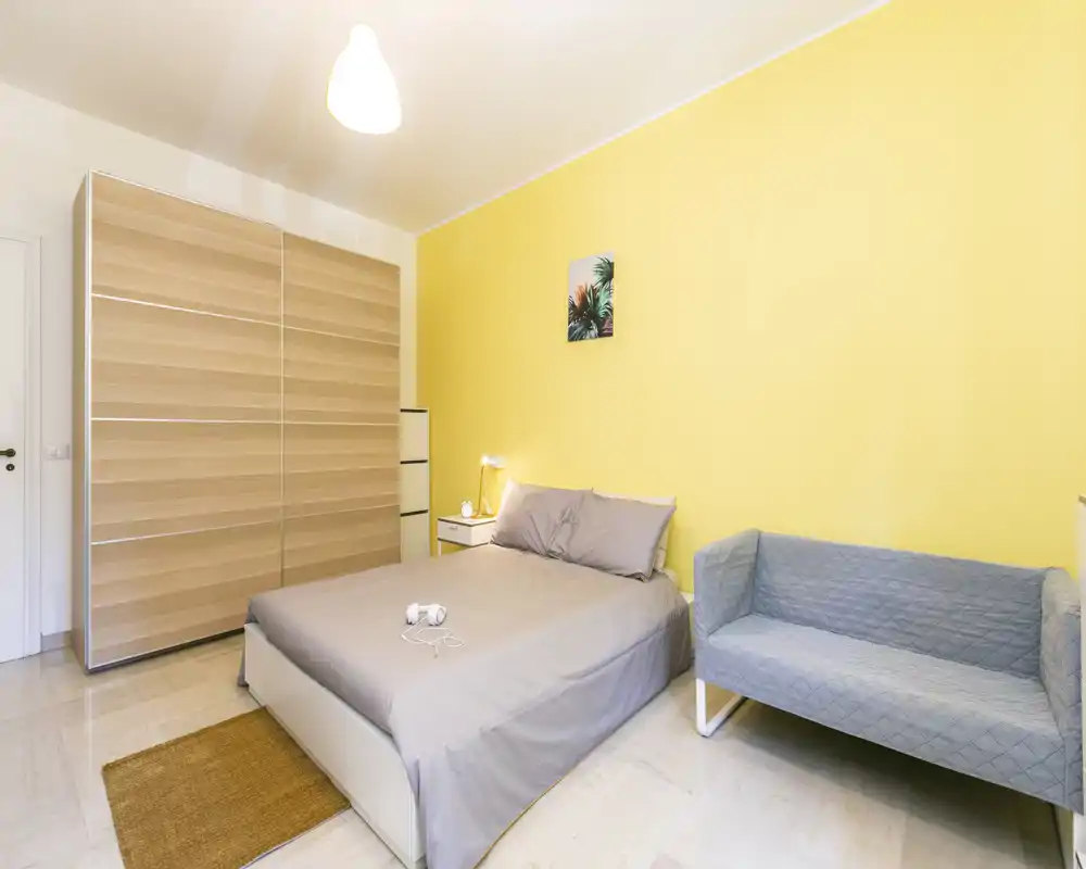 Dormitorio moderno y luminoso con cama doble, mesita de noche y un sofá compacto frente a una pared de acento amarilla — ideal para una estancia acogedora.