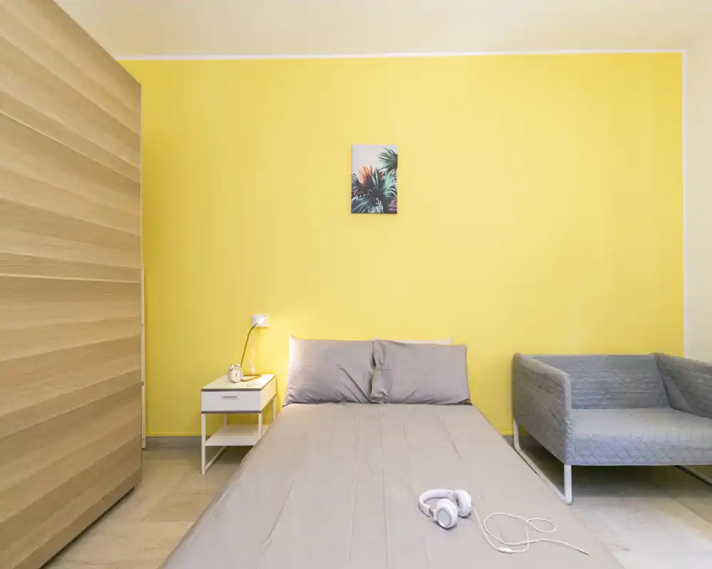 Dormitorio moderno y luminoso con cama doble bien hecha frente a una pared amarilla, mesita de noche con lámpara, armario y un sillón tapizado. Líneas limpias y decoración minimalista crean un espacio fresco y acogedor, perfecto para alquileres vacacionales.