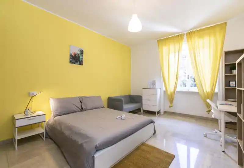 Camera da letto luminosa e ben arredata con letto matrimoniale contro una parete gialla, poltroncina, scrivania e luce naturale dalla finestra — accogliente e ariosa.