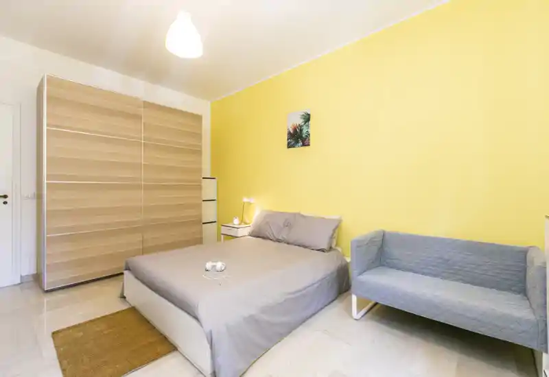 Camera da letto luminosa e moderna con letto matrimoniale, comodino e un piccolo divano contro una parete gialla vivace — perfetta per un soggiorno accogliente.