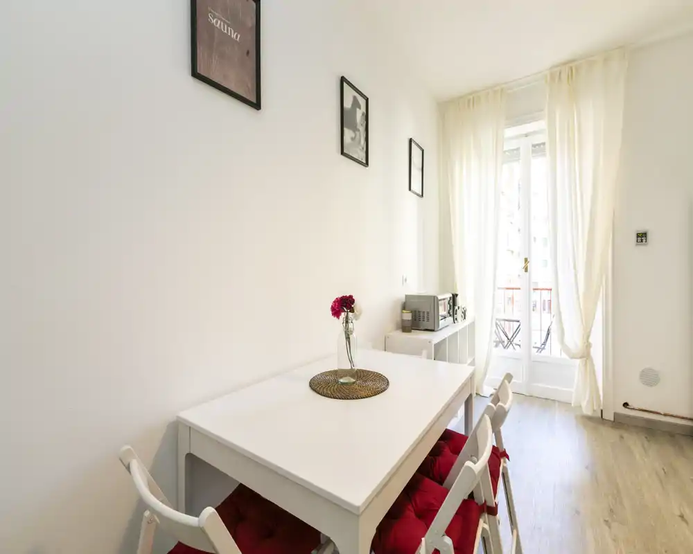 Zona de comedor luminosa y minimalista con mesa blanca, sillas con cojines rojos y un jarrón con flores junto a la puerta del balcón que deja entrar luz natural.
